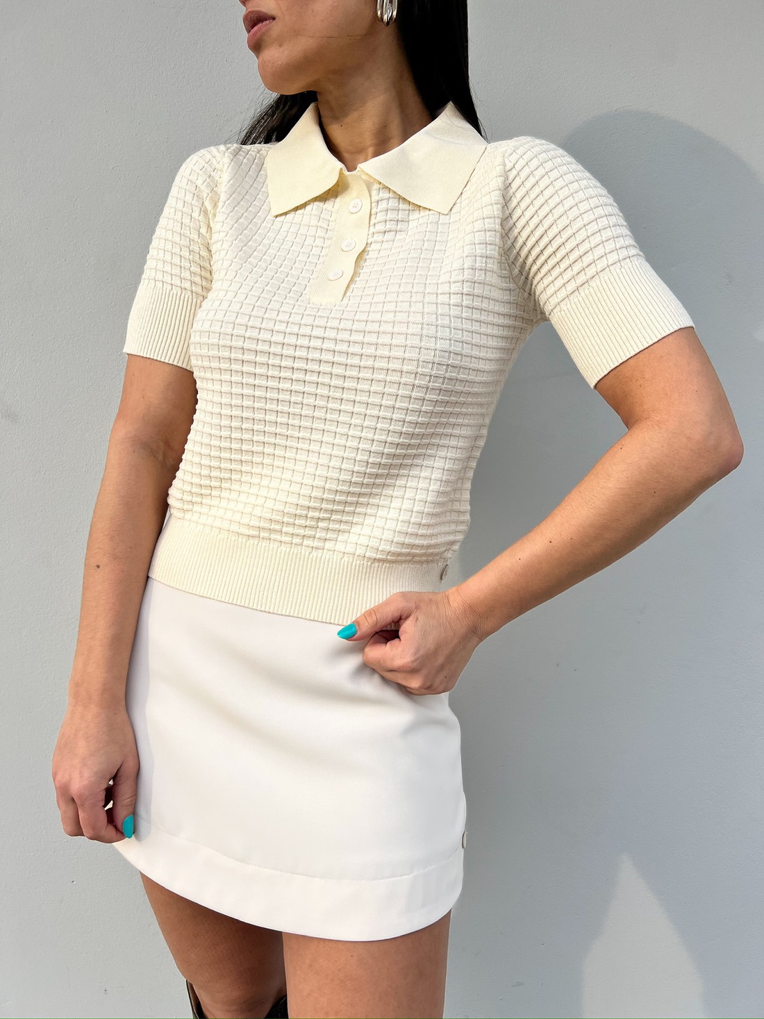 Blusa polo bege em tricô texturizado Blusa de tricô bege feminina com textura quadriculada, gola polo e botões, usada com saia branca curta.