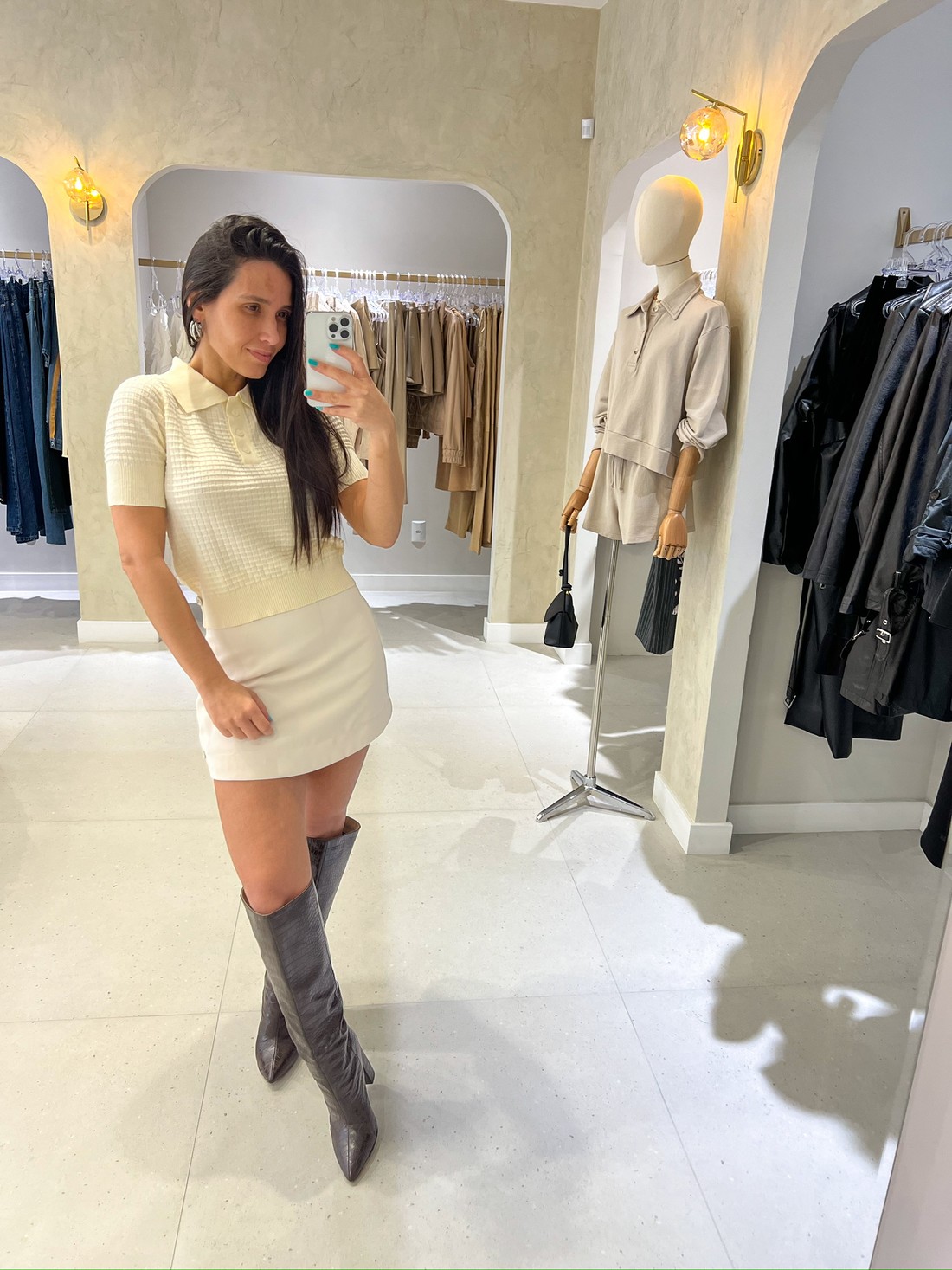 Look elegante com tricô bege e saia branca Look feminino com blusa de tricô bege de manga curta e gola polo, combinada com saia branca curta e bota cano alto marrom.
