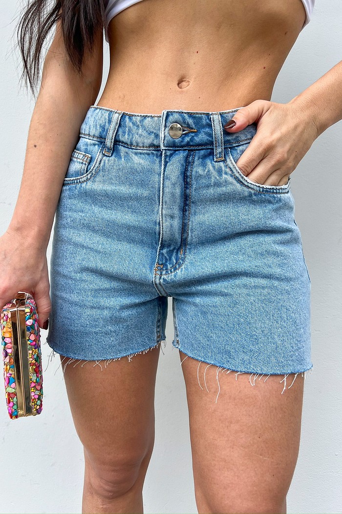 Shorts Jeans Bia MiniMoni em denim azul-claro, barra desfiada, vista frontal com mão no bolso, foco no caimento