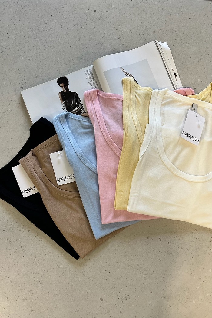 Opções de Cores da Regata Podrinha Essencial Todas as cores da Regata Básica Podrinha empilhadas ou lado a lado, mostrando a variedade de tons: Amarelo Manteiga, Branco, Rosa Ballet, Azul Céu e Preto.