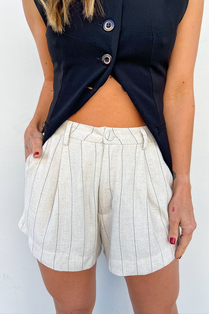 Modelo usando o Shorts de alfaiataria com risca de giz e um colete com uma proposta aberta, mostrando os detalhes do shorts, seu botão forrado e também a versatilidade da peça.